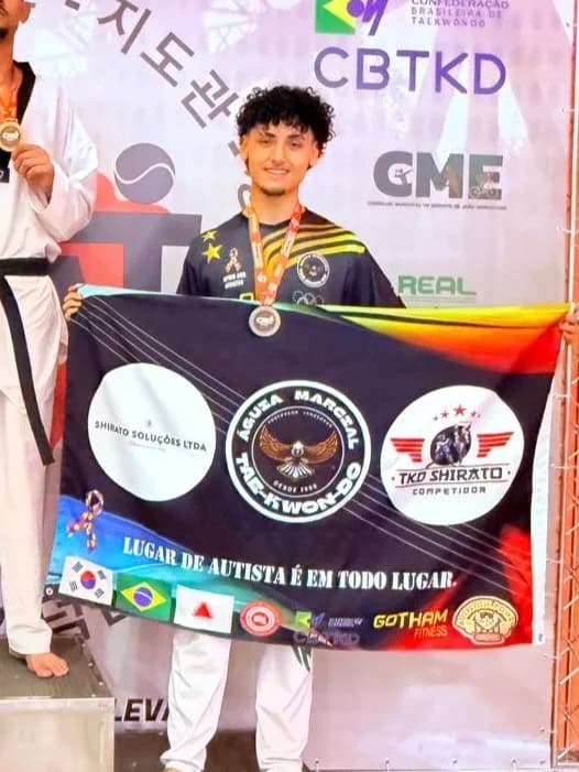 Atleta do TKD Shirato Competidor no pódio da CBTKD com a bandeira do Instituto, onde se lê Lugar de autista é em todo lugar