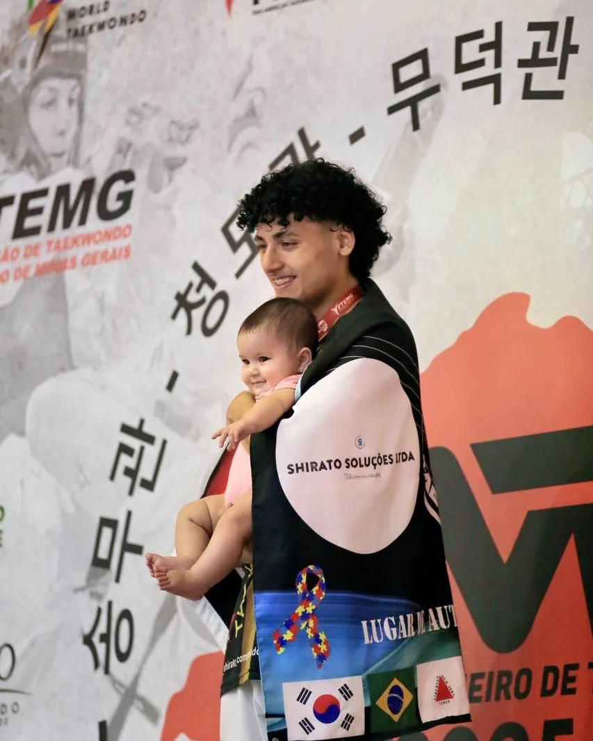 Christopher Shirato sorrindo no Campeonato Mineiro de 2025, com um bebê no colo e a bandeira do Instituto sobre os ombros