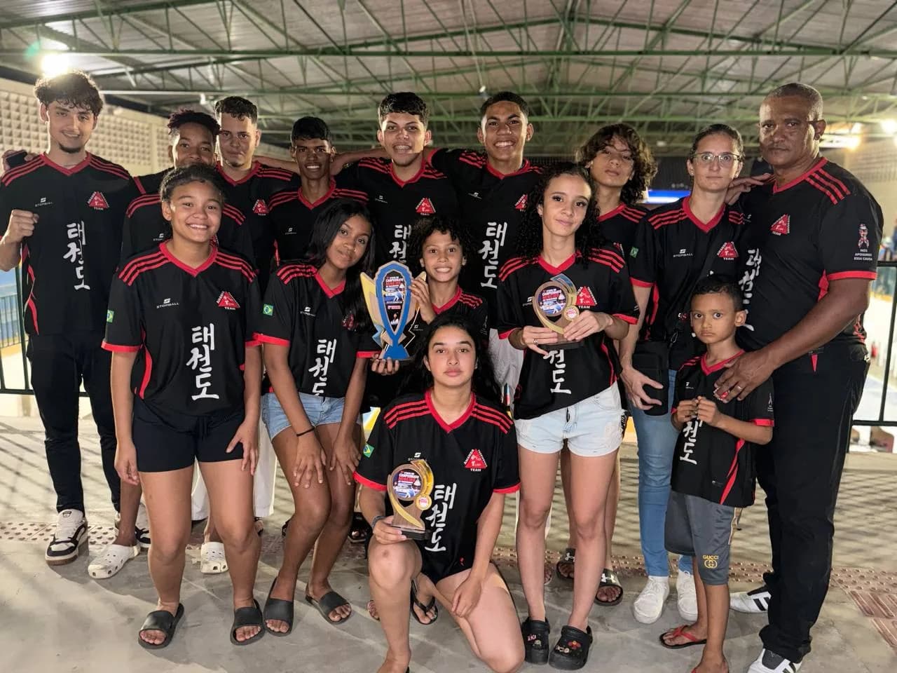 Atletas, professor e familiares reunidos com os troféus de competição, de camisa preta da equipe