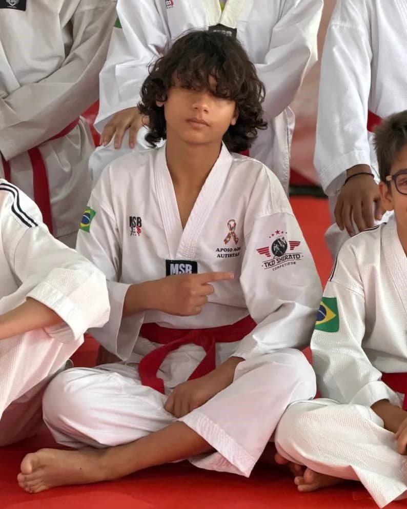 Kayky Shirato sentado no tatame com a turma, de dobok branco com o emblema de apoio aos autistas, apontando para o peito