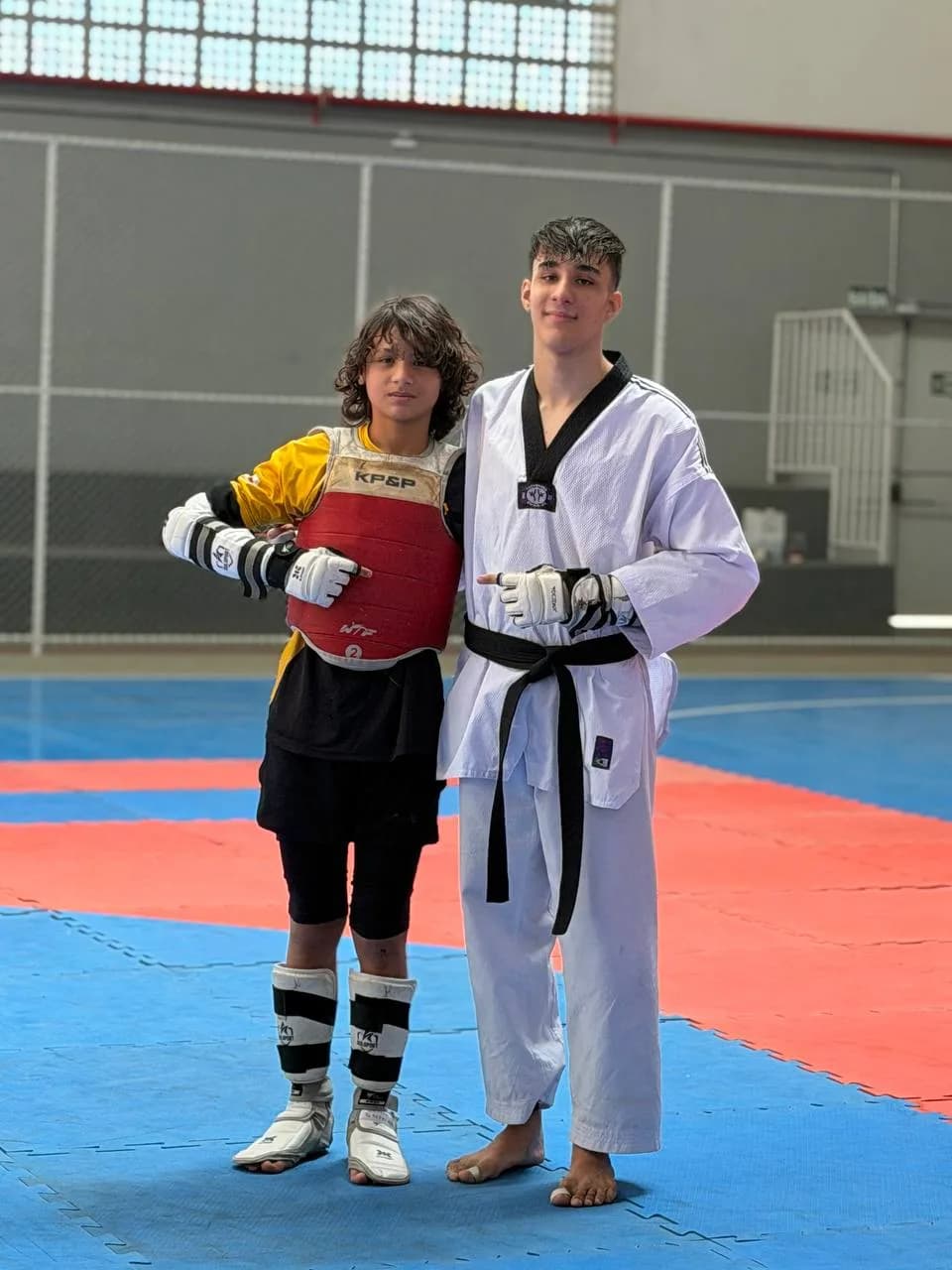 Kayky Shirato de protetor de tronco e luvas ao lado de atleta faixa preta, no tatame do ginásio de treino