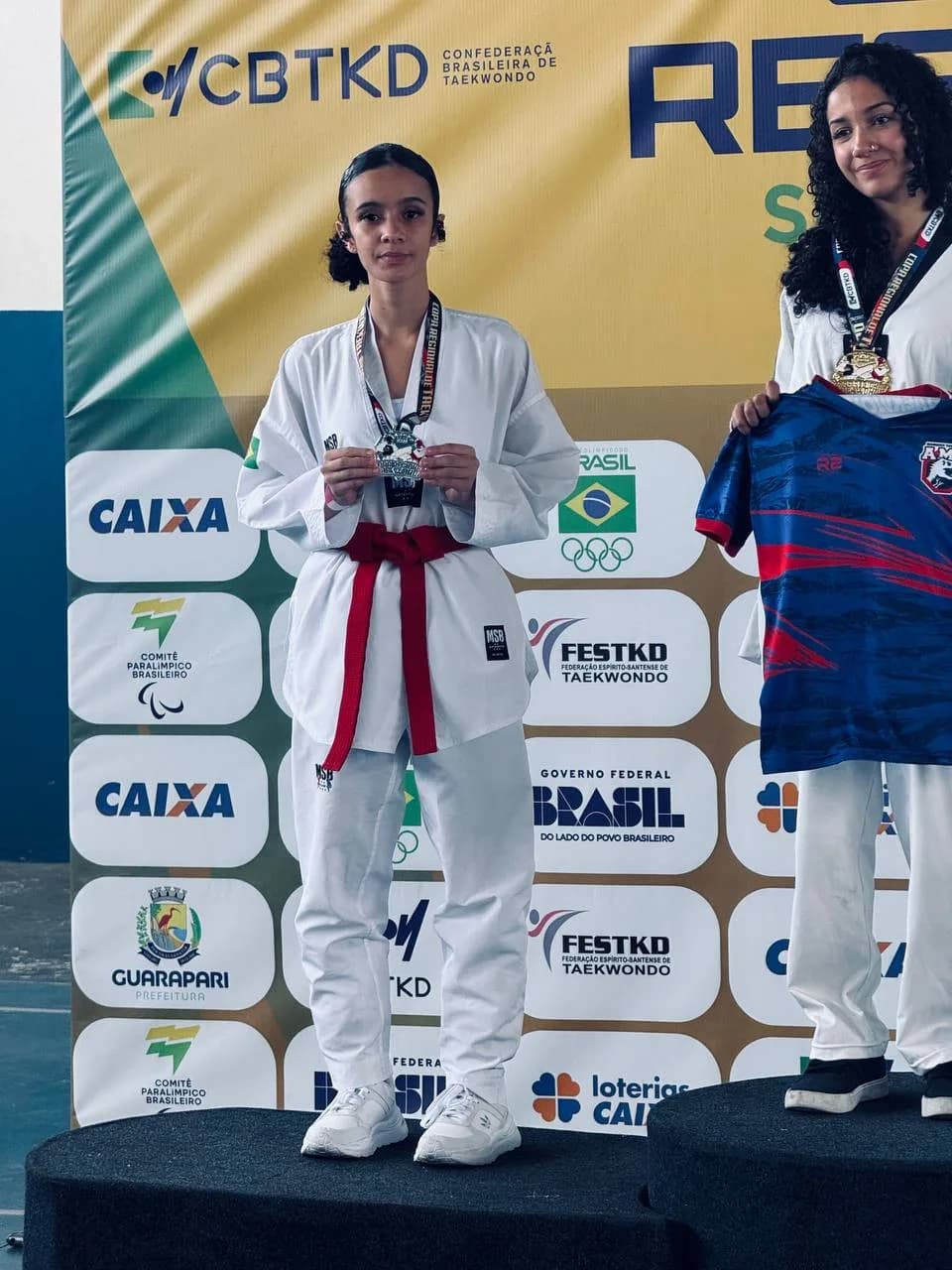 Atleta no pódio com a medalha de prata, diante do painel da Confederação Brasileira de Taekwondo