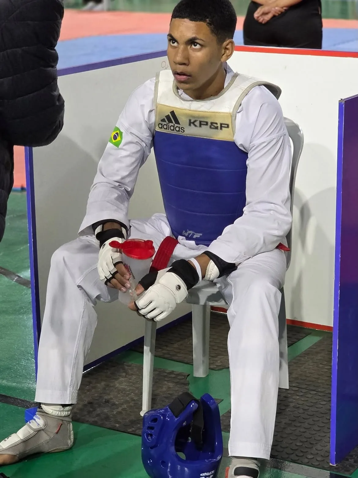 Lucas Lobo sentado à beira do tatame, de protetor de tronco azul, esperando a chamada para a luta